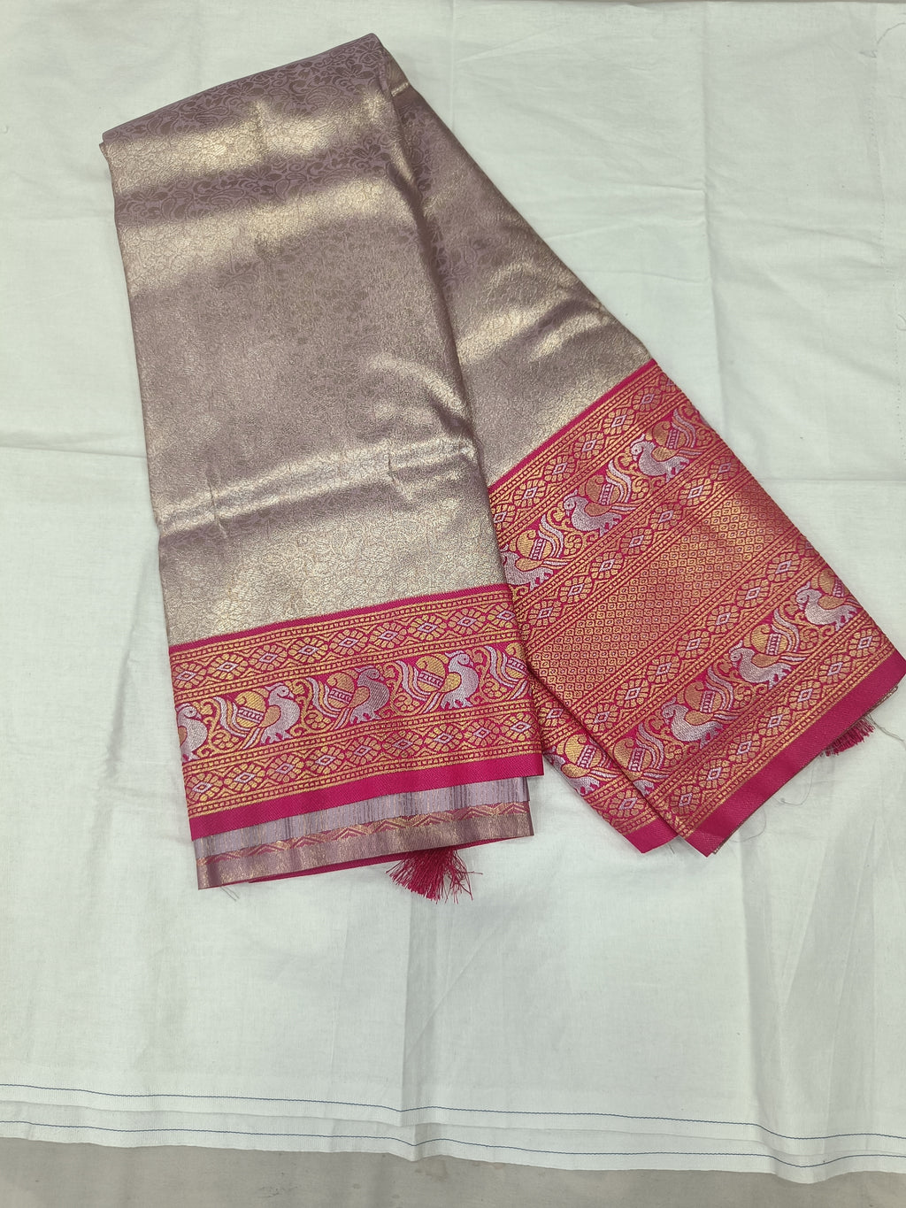 Folded light grey Banarasi silk saree with pink-gold border and matching blouse fabric