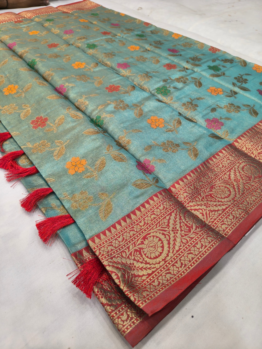 Sky-blue silk saree laid flat, floral motifs, red-gold border with tassels on edge